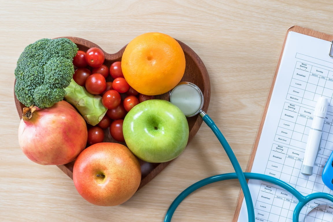 A heart-shaped bowl of fruits and vegetables, a stethoscope and a glucometer on a table with a blood sugar tracker.